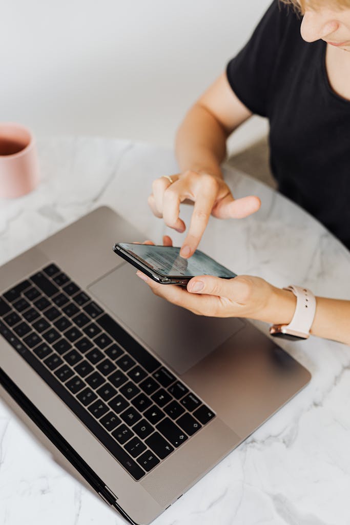 Adult using smartphone and laptop on a marble table for tech work.